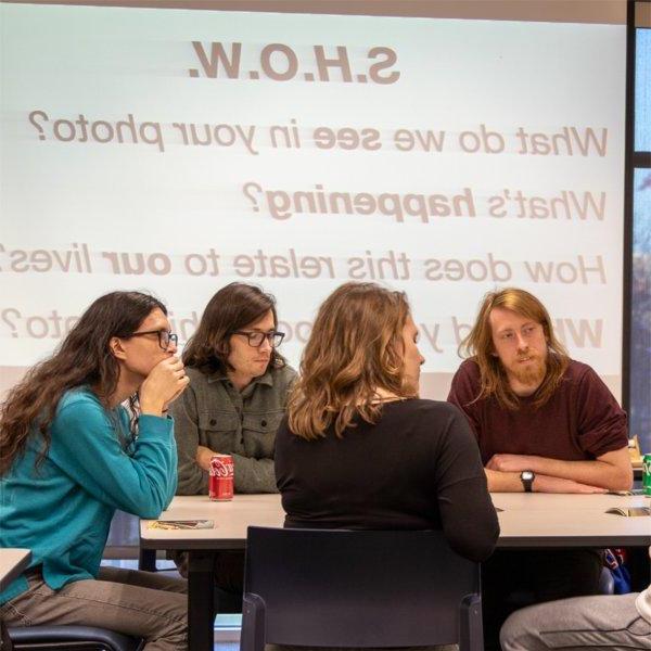 four students at table with projection on screen in background, asking SHOW, what do we see in your photo?